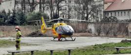 Na zdjęciu widzimy helikopter, który wylądował na placu o nawierzchni betonowej koloru szarego. Helikopter koloru żółtego z czerwonymi poziomymi pasami na bokach. Prawe drzwi maszyny są otwarte przy nich stoi ratownik ubrany w pomarańczowy kombinezon. Bliżej kadru widzimy stojącego strażaka, ubrany jest w ubranie specjalne koloru piaskowego, czerwony hełm, biała maska na twarzy, niebieskie rękawiczki. W tle widzimy krzewy bez liści, mur oraz budynki ( kamienice ) oraz część zabudowań kościoła.