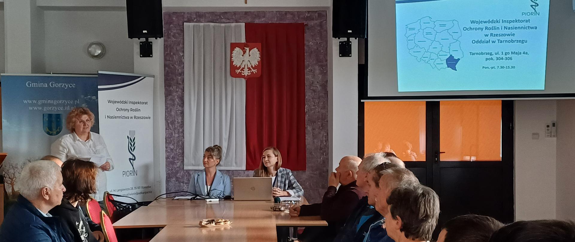 Grupa osób siedząca przy stole. Dwie kobiety siedzą przodem do grupy. Za nimi jest godło na biało czerwonej fladze. Po prawej stronie wyświetlana jest prezentacja na monitorze. Po prawej stronie stoi baner z logiem instytucji.