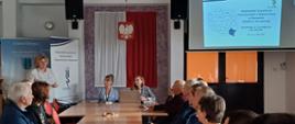 Grupa osób siedząca przy stole. Dwie kobiety siedzą przodem do grupy. Za nimi jest godło na biało czerwonej fladze. Po prawej stronie wyświetlana jest prezentacja na monitorze. Po prawej stronie stoi baner z logiem instytucji.