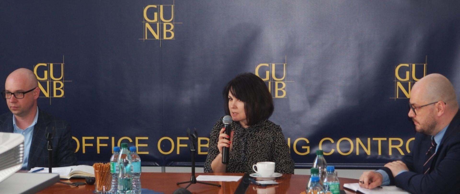 Trzy osoby siedzące przy stole konferencyjnym z mikrofonami i dokumentami; w tle baner GUNB – Office of Building Control.