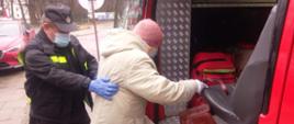 Strażak pomaga starszej kobiecie wejść do samochodu pożarniczego. Kobieta jest asekurowana przez strażaka, prawą ręką wspiera się o fotel w pojeździe. 