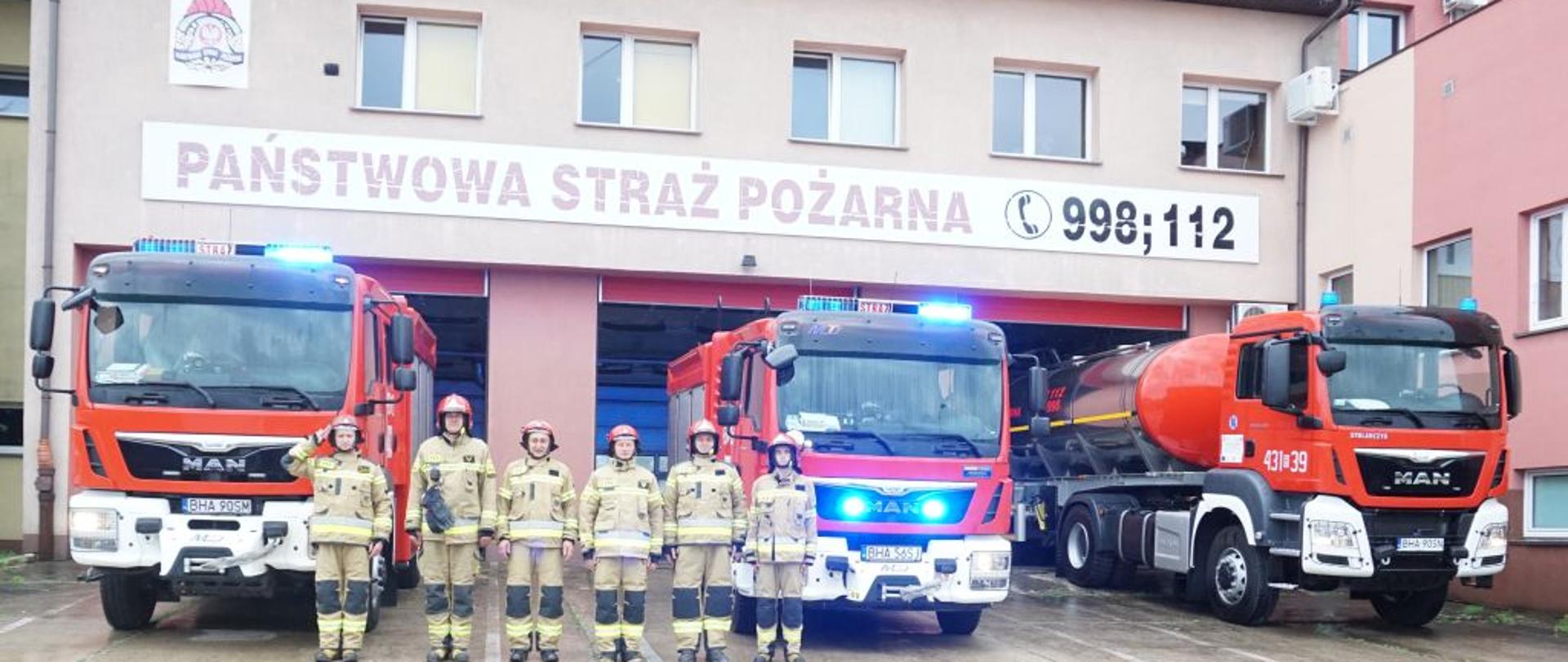 Na tle trzech czerwonych strażackich wozów bojowych stoi sześciu strażaków w piaskowych mundurach bojowych. W samochodach błyskają niebieskie sygnały świetlne.