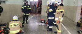 Zdjęcie przedstawia garaże Komendy Powiatowej Państwowej Straży Pożarnej w Radziejowie oraz strażaków OSP podczas wykonywania ćwiczenia z przecinarką do stali i betonu oraz piłą do cięcia drewna