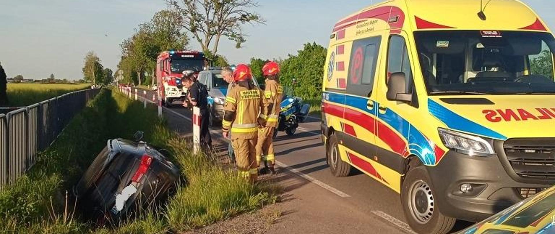 Na drodze z prawej strony stoi żółta karetka pogotowia za nią samochody strażackie. Z lewej strony w rowie leży samochód. Pogodnie.