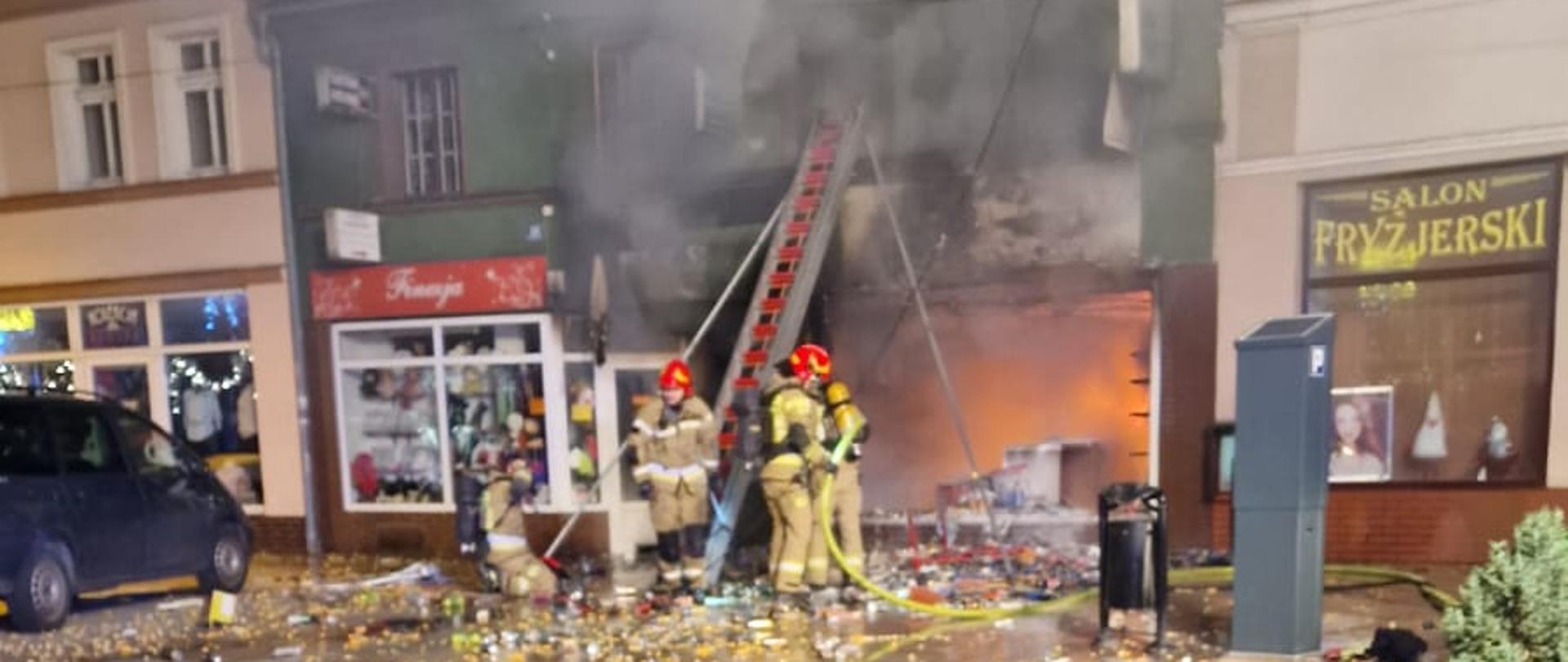 Cały lokal sklepu z alkoholem pali się, widać ogień. Strażacy prowadzą akcję gaśniczą.