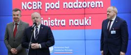 Na tle wbudowanego w ścianę wielkiego ekranu z napisem NCBR pod nadzorem Ministra Nauki stoi minister Wieczorek, obok niego wiceminister Gzik który mówi do mikrofonu na stojaku, obok nich z boku stoi mężczyzna w garniturze.