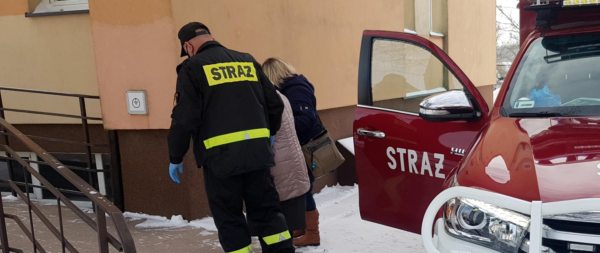 Na zdjęciu znajduje się samochód strażacki z otwartymi drzwiami a obok niego strażak prowadzący starszą osobę do punktu szczepień.