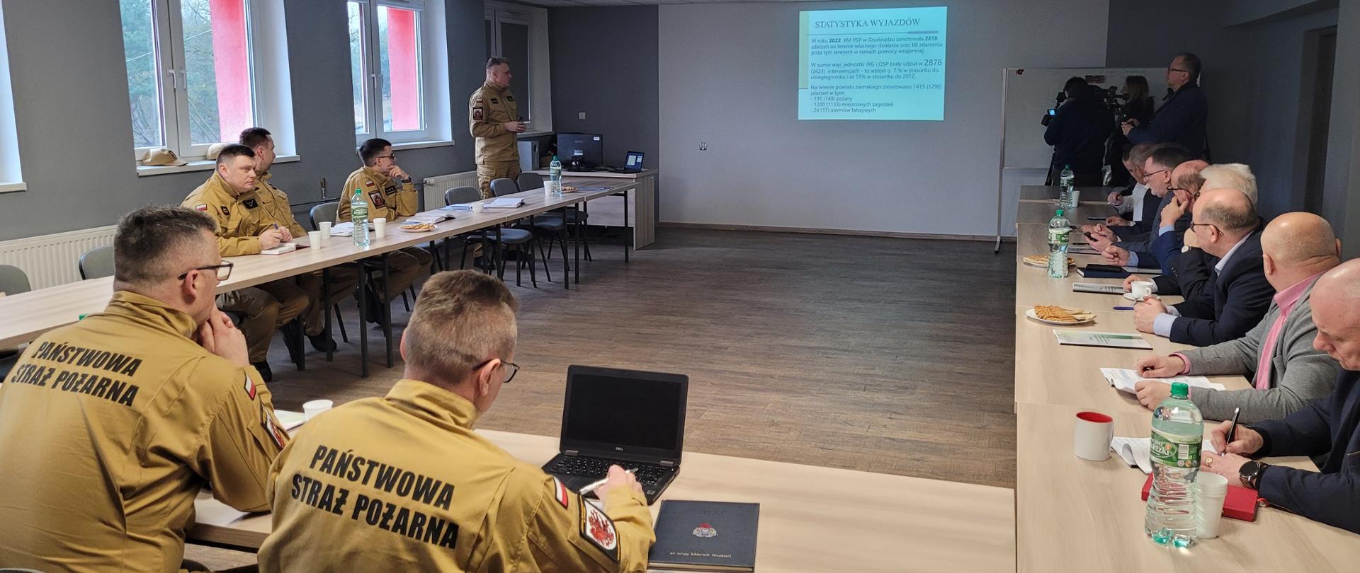 Sala wykładowa, w której przy stołach siedzą strażacy w piaskowych mundurach oraz osoby cywilne. Na jednej ze ścian wyświetlana jest prezentacja multimedialna.