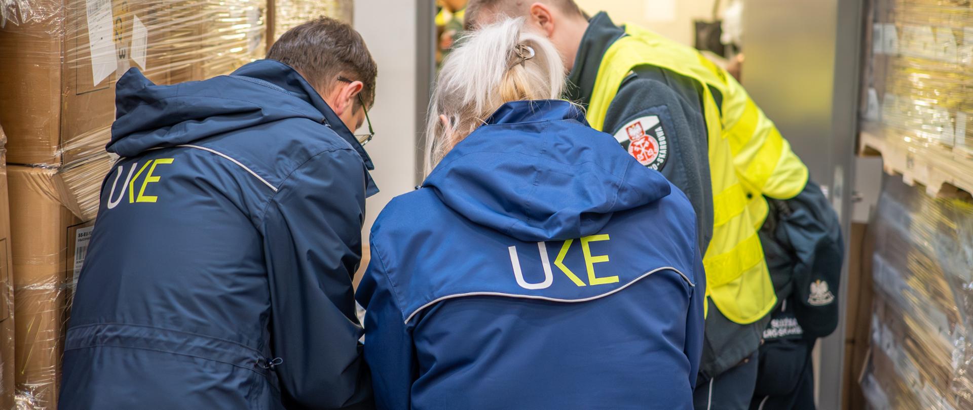 Trzy osoby w odzieży służbowej pochylają się nad stołem w magazynie, gdzie w tle znajdują się palety owinięte folią. Na plecach dwóch osób widoczny jest napis „UKE”, a obok stoi osoba w odblaskowej kamizelce.