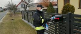 Na zdjęciu widać strażaka, który wrzuca ulotkę do skrzynki pocztowej, plot i część domu w tle droga trawnik i inny dom
