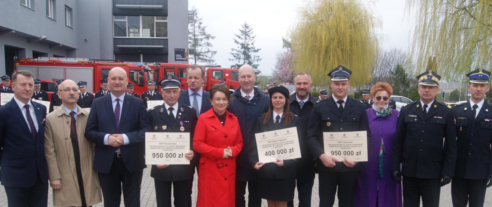 Grupa ludzi stoi do zdjęcia grupowego, wśród nich strażacy, w tle czerwony wóż strażacki i budynek 