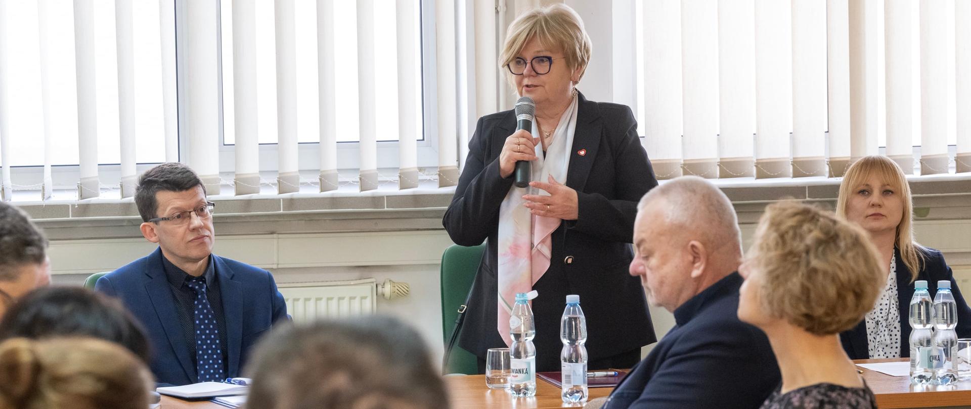 Stojąca kobieta z przemawia do zebranych osób, które siedzą przy stole konferencyjnym. Kobieta ma krótkie blond włosy jest ubrana w czarny żakiet a w ręku trzyma mikrofon. 