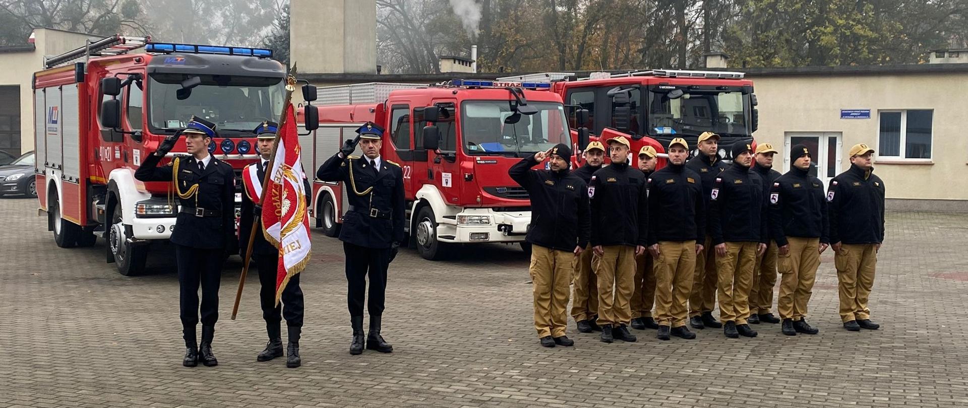 Zdjęcie przedstawia funkcjonariuszy KP PSP w Brodnicy podczas uroczystości upamiętniającej 105 rocznicę odzyskania niepodległości. Po lewo poczet flagowy. Za strażakami pojazdy gaśnicze JRG.