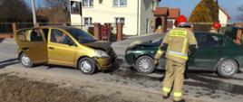 Zdjęcie przedstawia wypadek dwóch samochodów osobowych: Daewo Matiz oraz Opel Astra. Przed uszkodzonymi pojazdami stoi strażak. Na jezdni widać wycieki płynów eksploatacyjnych. W tle znajduje się wysoki budynek mieszkalny.
