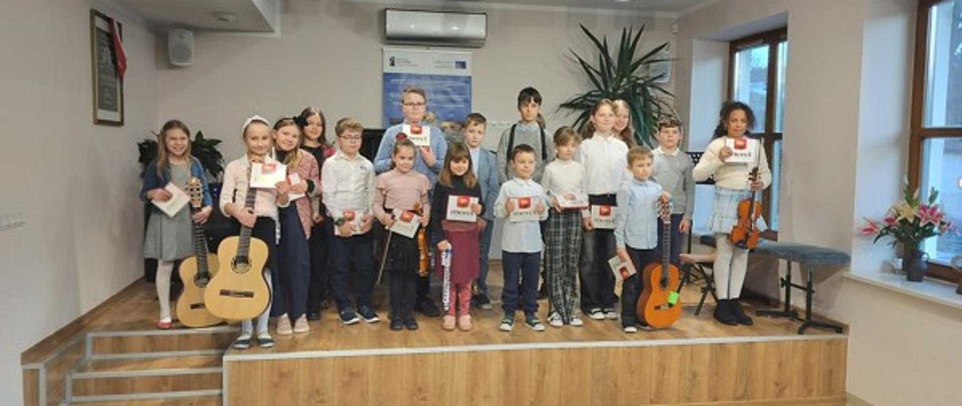 Fotografia przedstawiająca scenę we wnętrzu auli Państwowej Szkoły Muzycznej I i II stopnia w Nowym Targu, Filia w Jaworkach. Na scenie stojące dzieci ze swoimi instrumentami muzycznymi biorące udział w Karnawałowej Audycji Muzycznej. Na pierwszym planie publiczność. Za dziećmi fortepian oraz baner unijny, na suficie zapalone lampy. Z prawej strony dwa okna.