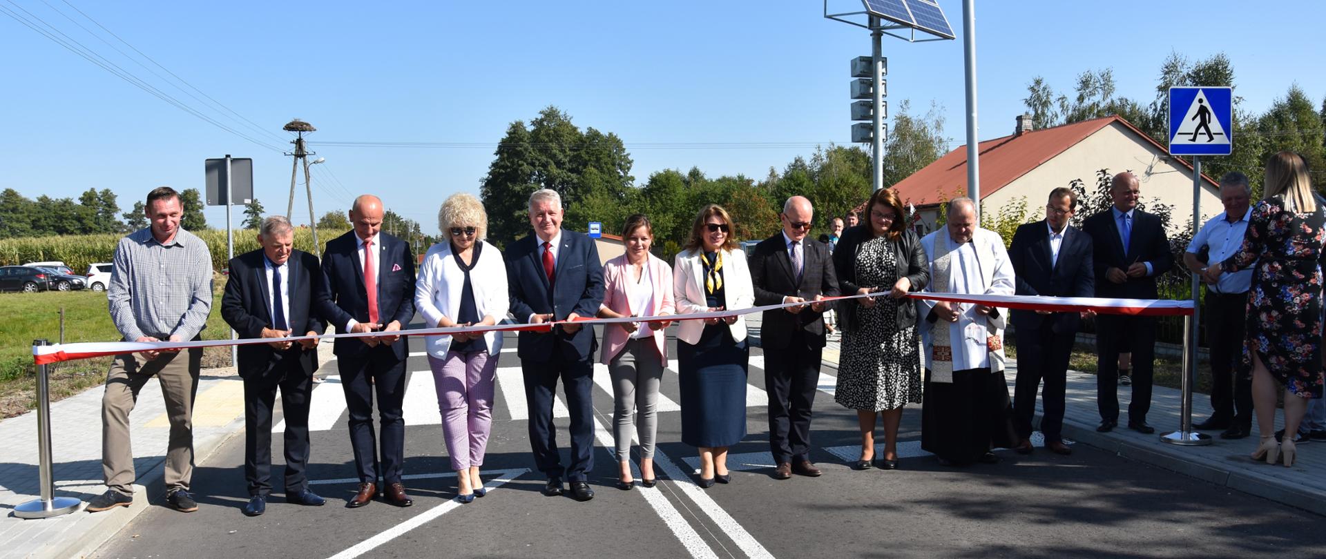 Na pierwszym planie znajduje się dwanaście elegancko ubranych osób, które stoją na jezdni zmodernizowanej drogi, przed biało-czerwoną wstęgą. W tle kępa drzew, obiekty inżynierii drogowej (między innymi znaki drogowe) i piękne błękitne niebo. Uroczystości towarzyszyła piękna, słoneczna pogoda