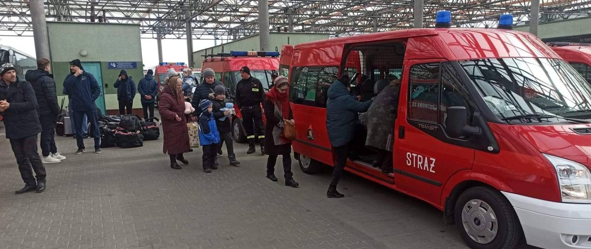 Do busów strażackich koloru czerwonego wsiadają ludzie, kobiety z dziećmi