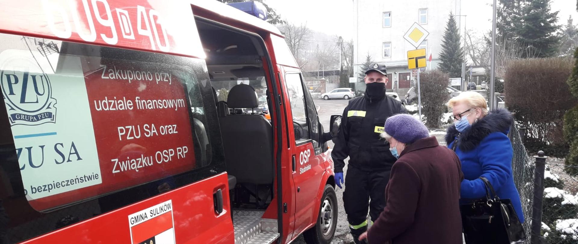 Samochód typu bus należący do OSP z numerem operacyjnym 609D40. Otwarte drzwi boczne na których znajduje się herb gminy Sulików. Dwie kobiety wsiadają do pojazdu. Obok stoi strażak OSP.