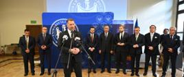 Na dużej oświetlonej przez wpadające przez duże okna światło sali pod niebieską ścianką stoi w szeregu kilku mężczyzn w garniturach, przed nimi minister Czarnek stoi i mówi do kilku mikrofonów na stojakach.