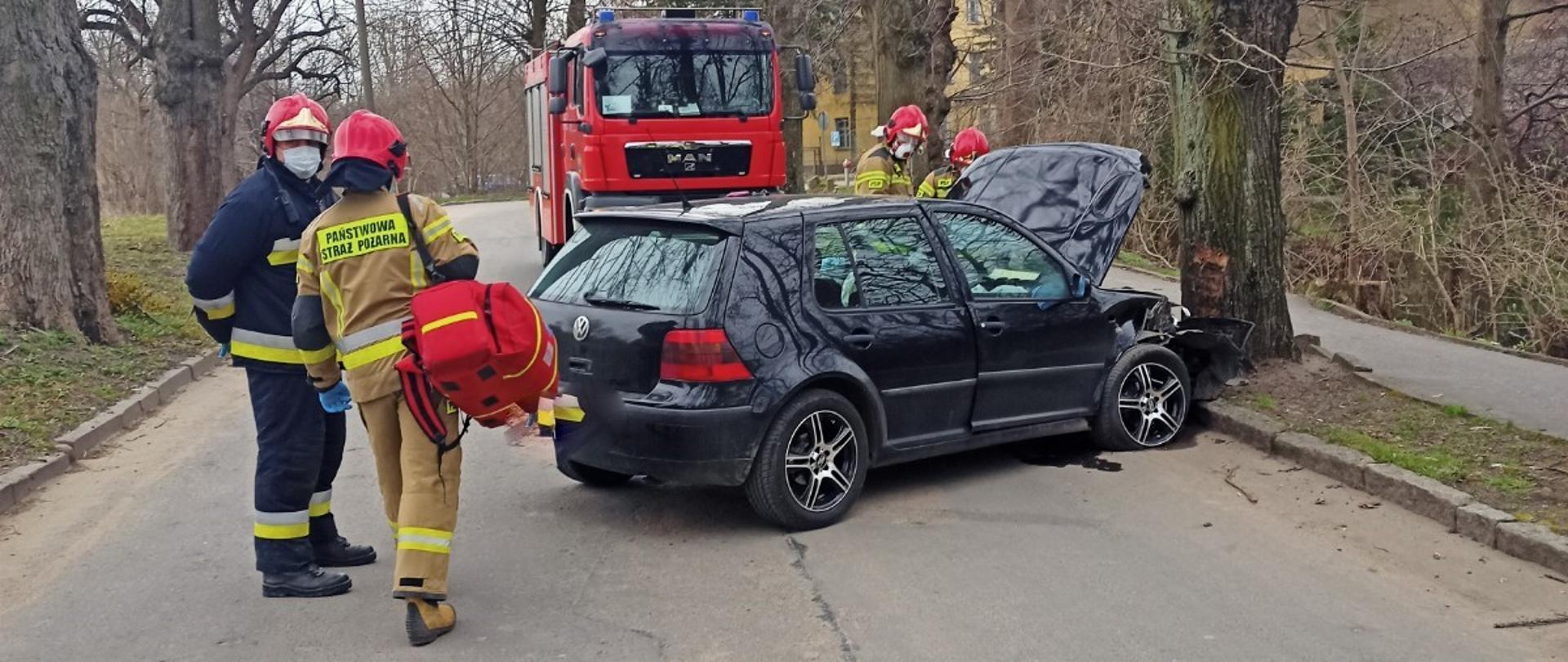 Zdjęcie przedstawia strażaków skupionych przy samochodzie osobowym, który uderzył czołowo w przydrożne drzewo. Jeden ze strażaków niesie torbę medyczną R1 do samochodu pożarniczego znajdującego się na drugim planie.