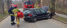 Zdjęcie przedstawia strażaków skupionych przy samochodzie osobowym, który uderzył czołowo w przydrożne drzewo. Jeden ze strażaków niesie torbę medyczną R1 do samochodu pożarniczego znajdującego się na drugim planie.