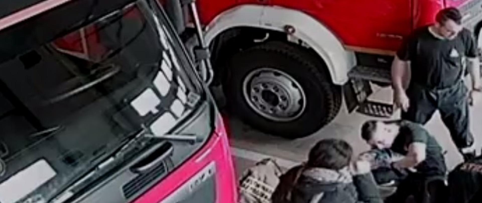 Zdjęcie obrazuje strażaków i inną osobę oraz pojazdy pożarnicze