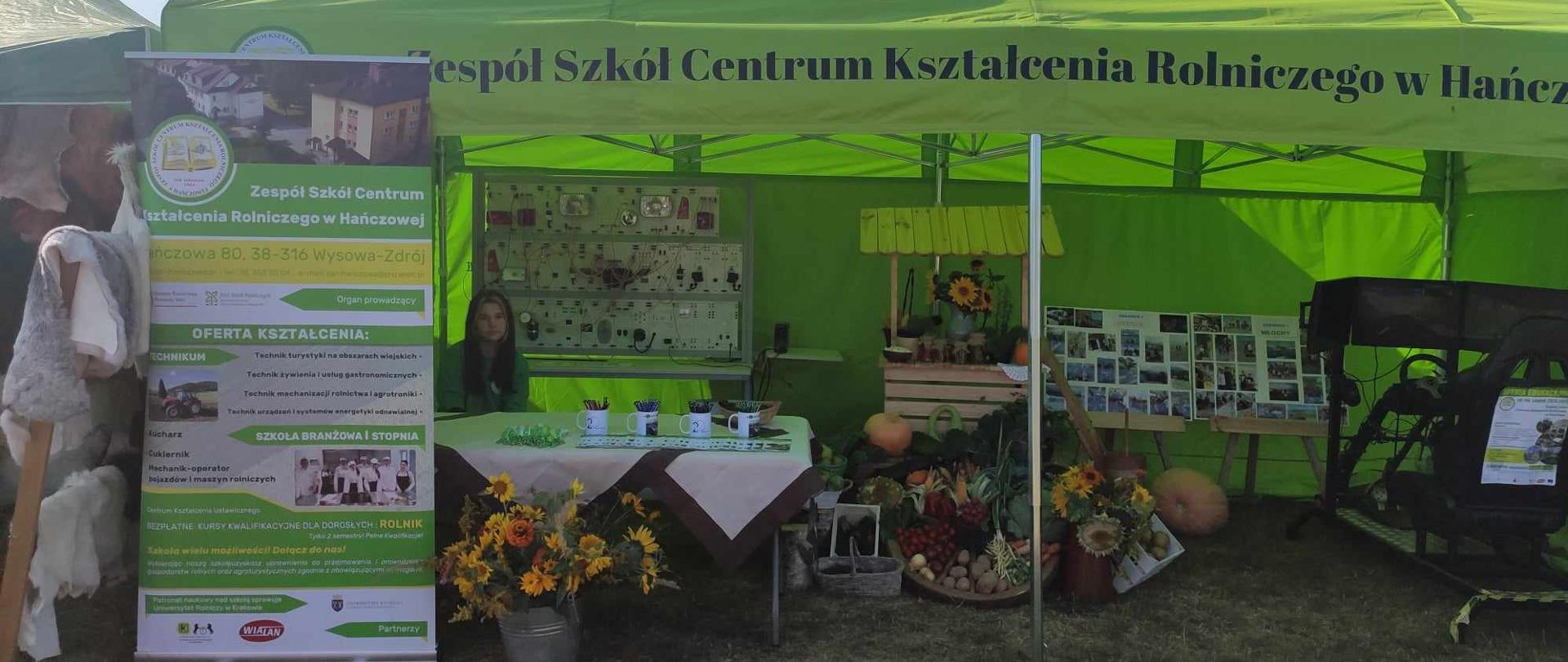 Widok na front zielonego namiotu wystawowego Zespołu Szkół Centrum Kształcenia Rolniczego w Hańczowej. Wewnątrz widoczne stoisko z warzywami, tablice edukacyjne i symulator jazdy.