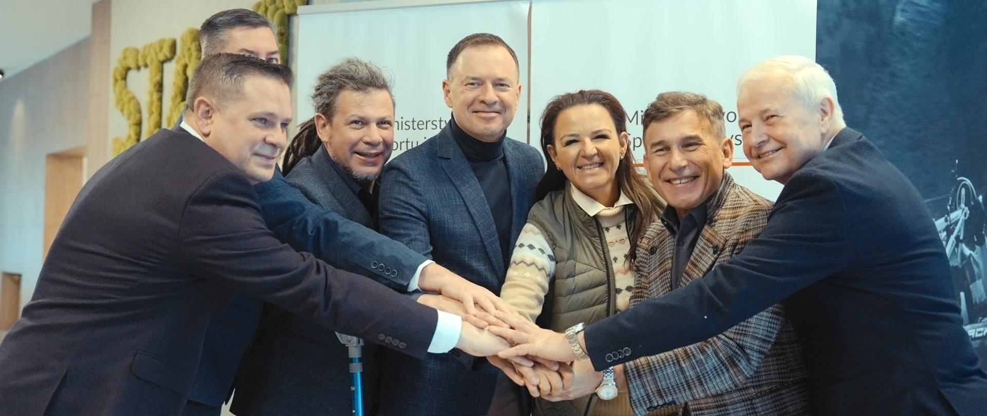 Konsultacje nt. strategii sportu w Wiśle. Wiceminister Piotr Borys wraz ekspertami trzymający się za ręce na tle ścianki z logiem ministerstwa