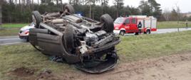 Zdjęcie przedstawia na pierwszym planie samochód osobowy na poboczu drogi wywrócony na dach, z rozbitym przodem. Na drugim planie za samochodem, na drodze asfaltowej stoi samochód zespołu ratownictwa medycznego oraz po prawej stronie lekki samochód gaśniczy Państwowej Straży Pożarnej. Na dalszym planie widzimy dwóch strażaków Państwowej Straży Pożarnej, jeden stoi na drodze asfaltowej, drugi przy samochodzie gaśniczym.