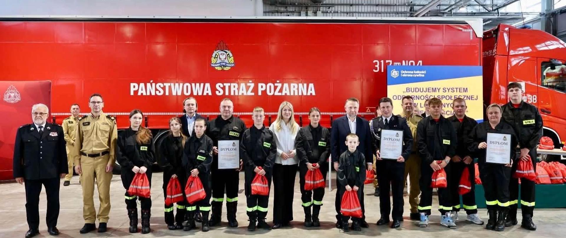 Szerokie ujęcie grupowe wykonane w przestronnej hali garażowej jednostki ratowniczo-gaśniczej. W tle stoi duży, czerwony wóz strażacki z białym napisem „PAŃSTWOWA STRAŻ POŻARNA” oraz plansza z nazwą konkursu: „BUDUJEMY SYSTEM ODPORNOŚCI SPOŁECZNEJ W LOKALNYCH SPOŁECZNOŚCIACH”. W jednym rzędzie stoi kilkanaście osób: umundurowani strażacy PSP w beżowych i ciemnych mundurach galowych, członkowie Młodzieżowych i Dziecięcych Drużyn Pożarniczych w czarnych mundurach koszarowych ze seledynowymi odblaskami i napisem „STRAŻ”, a także zaproszeni goście w cywilnych strojach wizytowych. Część uczestników trzyma w dłoniach oprawione dyplomy oraz małe, czerwone worki z upominkami.