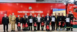 Szerokie ujęcie grupowe wykonane w przestronnej hali garażowej jednostki ratowniczo-gaśniczej. W tle stoi duży, czerwony wóz strażacki z białym napisem „PAŃSTWOWA STRAŻ POŻARNA” oraz plansza z nazwą konkursu: „BUDUJEMY SYSTEM ODPORNOŚCI SPOŁECZNEJ W LOKALNYCH SPOŁECZNOŚCIACH”. W jednym rzędzie stoi kilkanaście osób: umundurowani strażacy PSP w beżowych i ciemnych mundurach galowych, członkowie Młodzieżowych i Dziecięcych Drużyn Pożarniczych w czarnych mundurach koszarowych ze seledynowymi odblaskami i napisem „STRAŻ”, a także zaproszeni goście w cywilnych strojach wizytowych. Część uczestników trzyma w dłoniach oprawione dyplomy oraz małe, czerwone worki z upominkami.