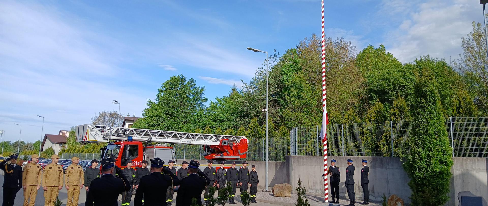 Strażacy ustawieni na placu wewnętrznym Komendy Miejskiej PSP w Mysłowicach na uroczystej zbiórce oddają hołd Fladze Rzeczpospolitej Polskiej wciąganej na maszt przez poczet flagowy przy odgrywanym hymnie państwowym.