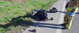 Uszkodzony motorower leży na poboczu drogi, obok urwane koło. Z boku stoi strażak, a z tyłu samochód strażacki.