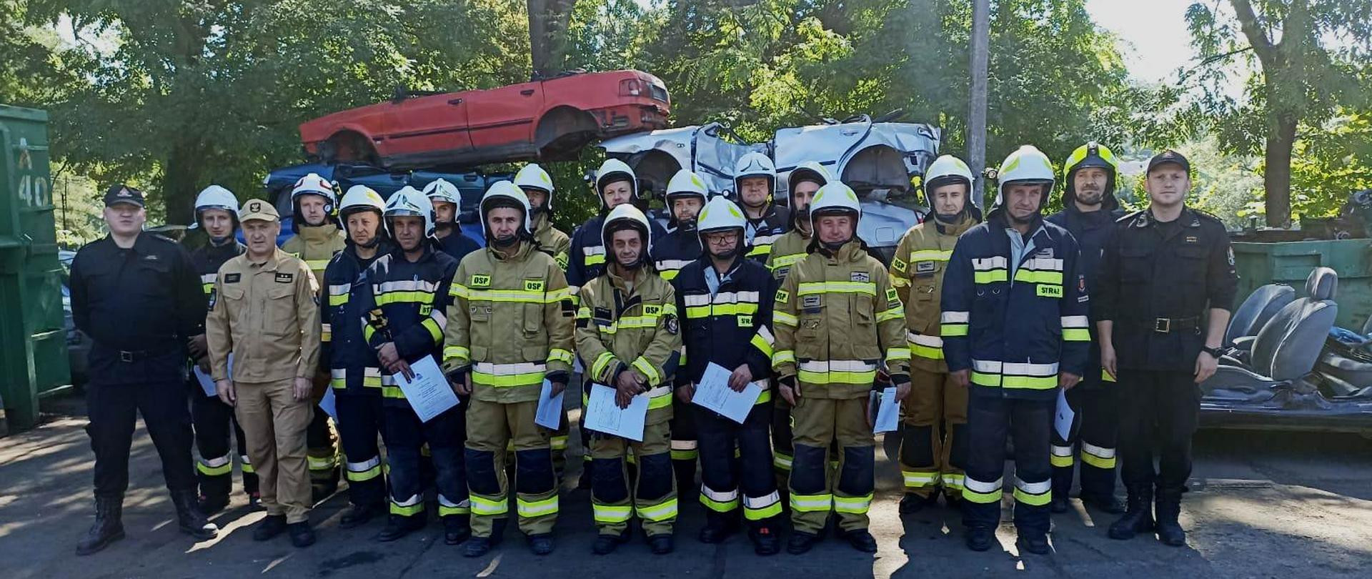 Kolorowa fotografia wykonana na zewnątrz, przedstawia strażaków ochotniczych straży pożarnych stojących w ubraniach specjalnych i hełmach oraz strażaków Państwowej Straży Pożarnej w jasnym i dowódczo-sztabowym ubraniu służbowym. 