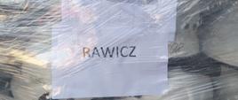 Sprzęt pożarniczy dla ukraińskich strażaków. Na europalecie są ułożone hełmy strażackie koloru białego. Hełmy są owinięte folią. Na folii przyklejone są dwie kartki: jedna z napisem "RAWICZ", druga to "Karta załadunkowa". 