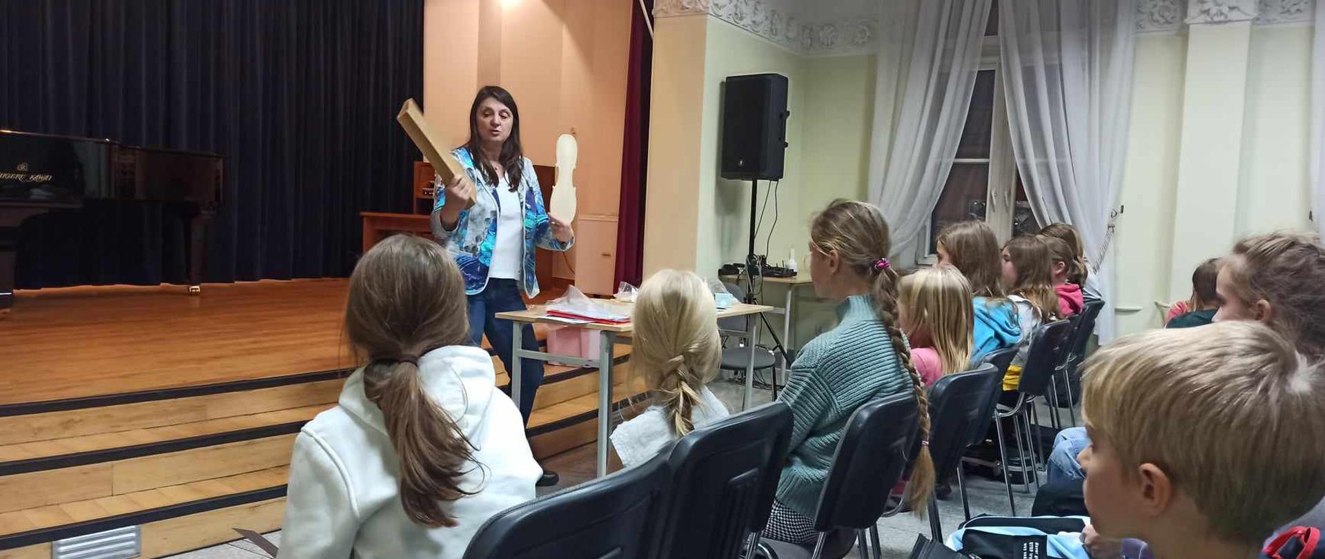 Fotogaleria z warsztatów lutniczych. Na zdjęciu lutnik pokazuje jak powstają skrzypce, jakie materiały służą do konserwacji instrumentu. Na sali jest dużo obserwatorów - uczniów szkoły muzycznej.