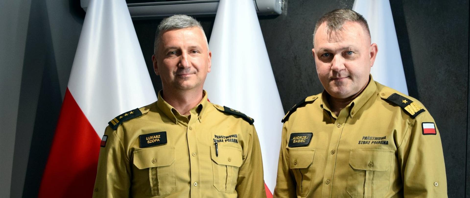 Zdjęcie zrobione wewnątrz pomieszczenia. Na zdjęciu dwóch oficerów Państwowej Straży Pożarnej. Za nimi ustawione są biało-czerwone flagi.