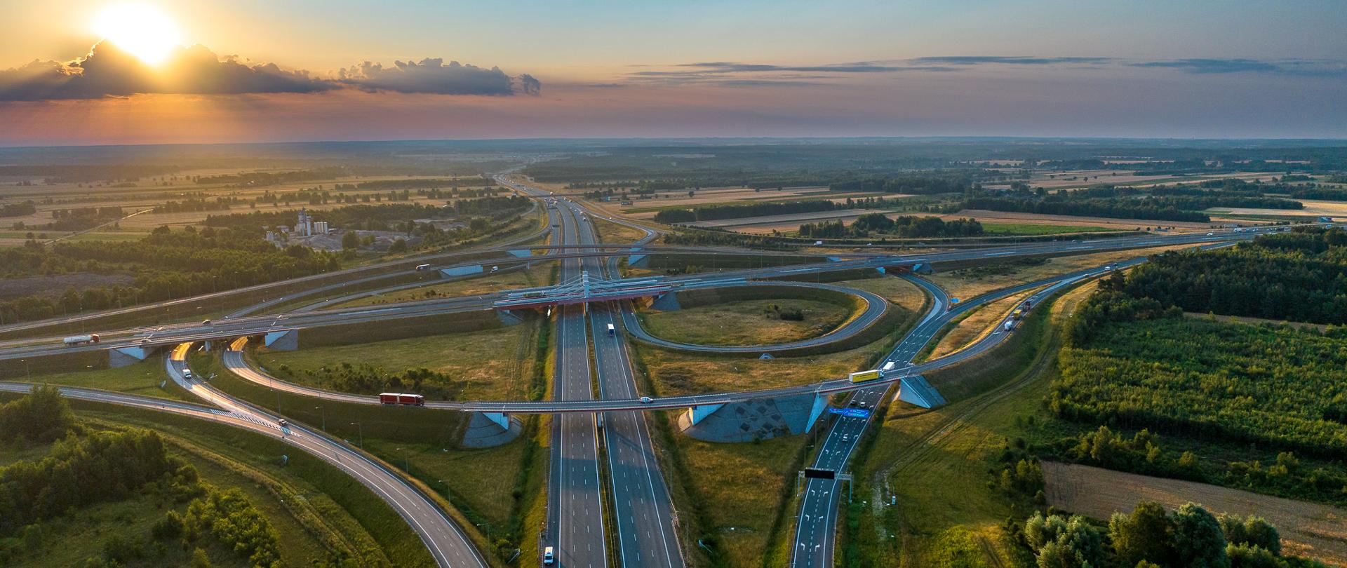 Widok z lotu ptaka na rozległy węzeł autostradowy o zachodzie słońca. Wielopoziomowe estakady i łącznice łączą główne trasy przebiegające przez otwarte tereny rolnicze i leśne.