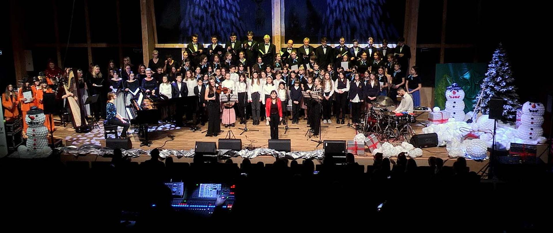 Duża grupa osób stojąca na scenie, wszyscy zwróceni twarzami w kierunku robiącego zdjęcie, w dole widać ciemne miejsca dla publiczności. Na scenie, po bokach, przed stojącymi osobami stoi fortepian i zestaw perkusyjny, po prawej stronie dekoracje przedstawiające śnieżne bałwany.