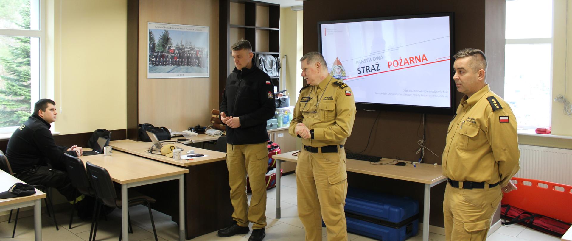 Trzech funkcjonariuszy Państwowej Straży Pożarnej stoi przed ekranem multimedialnym w sali szkoleniowej. Na ekranie wyświetlony jest logotyp PSP.