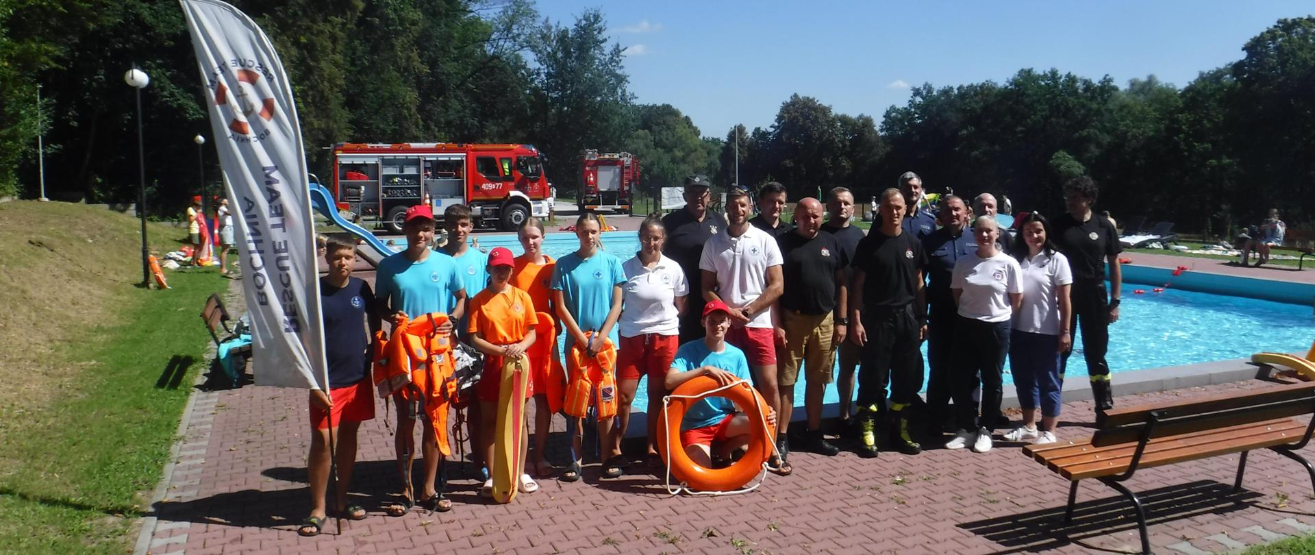 Zdjęcie przedstawia służby i inspekcję biorące udział w wydarzeniu. Na tle trawy i drzew znajduje się samochód strażacki. Na pierwszym planie widać przedstawicieli służb, dzieci oraz banery służb i inspekcji
