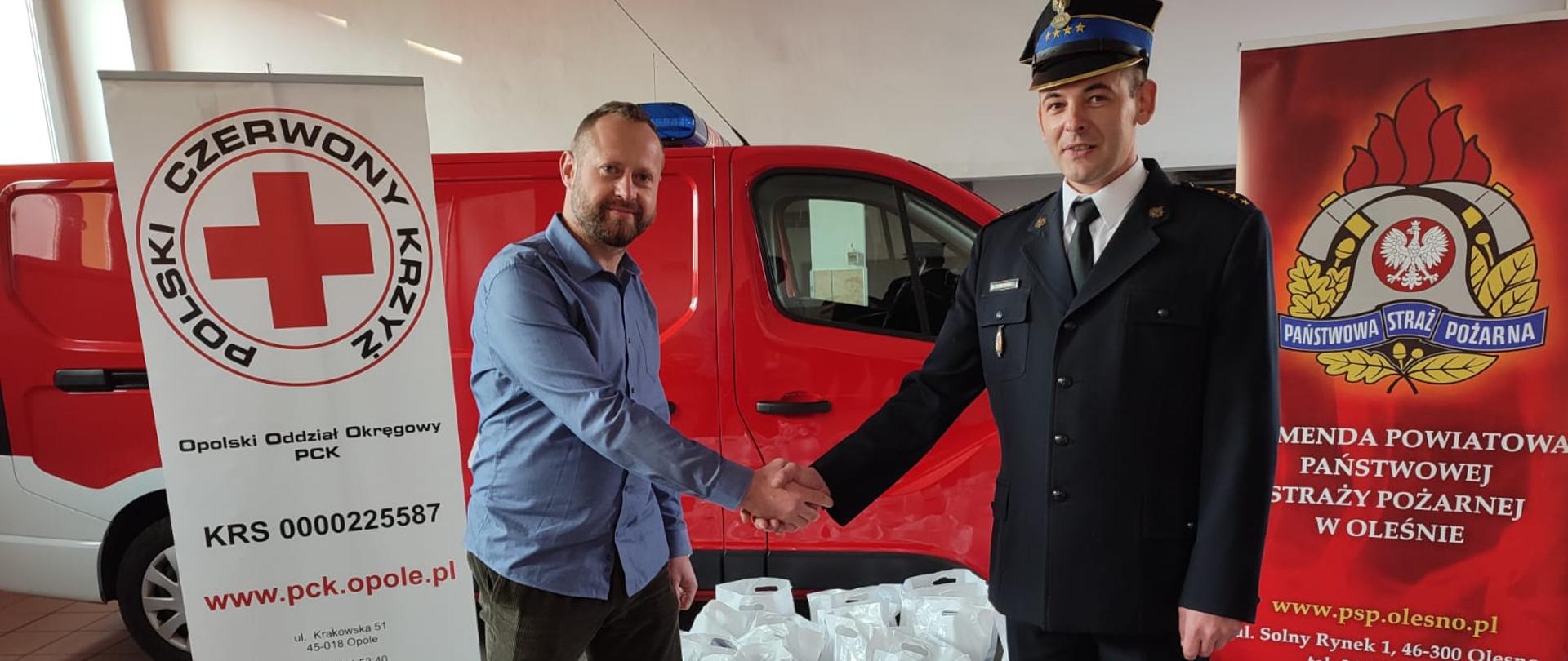 Strażak ubrany w mundur wyjściowy podaje prawą dłoń przedstawicielowi PCK. W tle samochód pożarniczy, banery PCK i PSP oraz podarunki w białych reklamówkach.