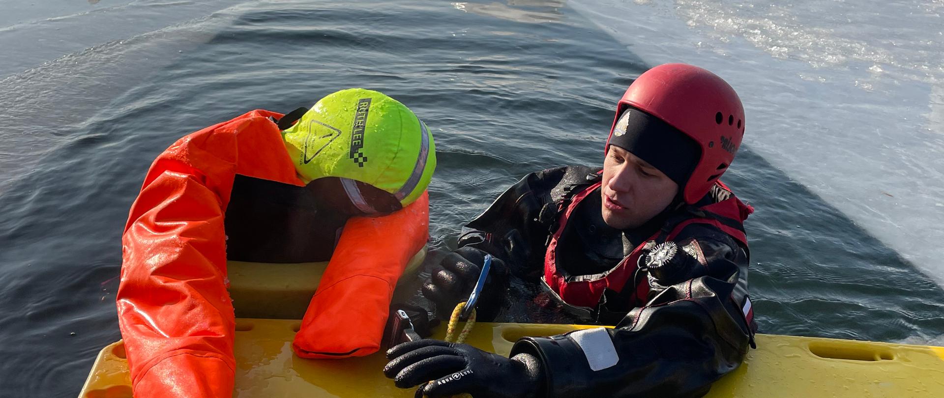 Na zdjęciu widoczny ratownik i manekin w przeręblu. Ratownik opiera się o żółtą deskę.