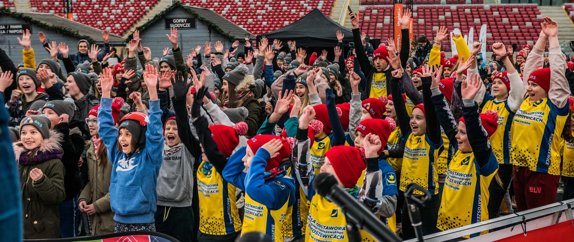 Tłum uśmiechniętych dzieci w czapkach zimowych z uniesionymi rękami wiwatuje podczas konferencji