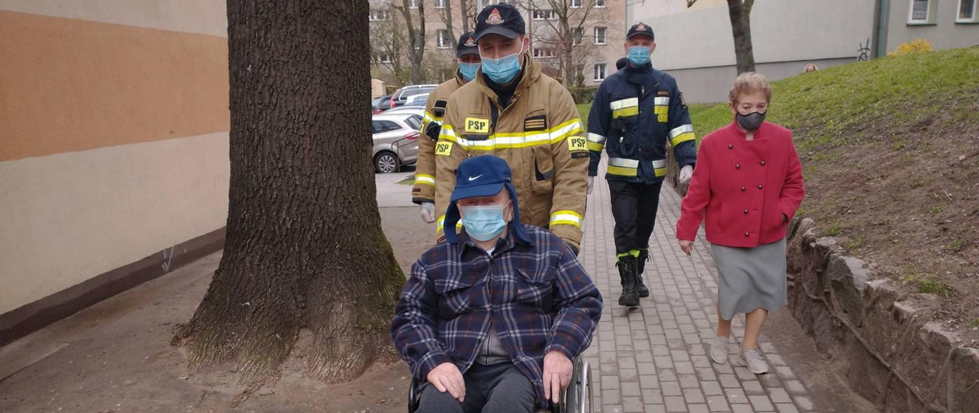 Na zdjęciu widać mężczyznę na wózku inwalidzkim prowadzonego i asystowanego przez trzech strażaków oraz kobietę, w tle bloki