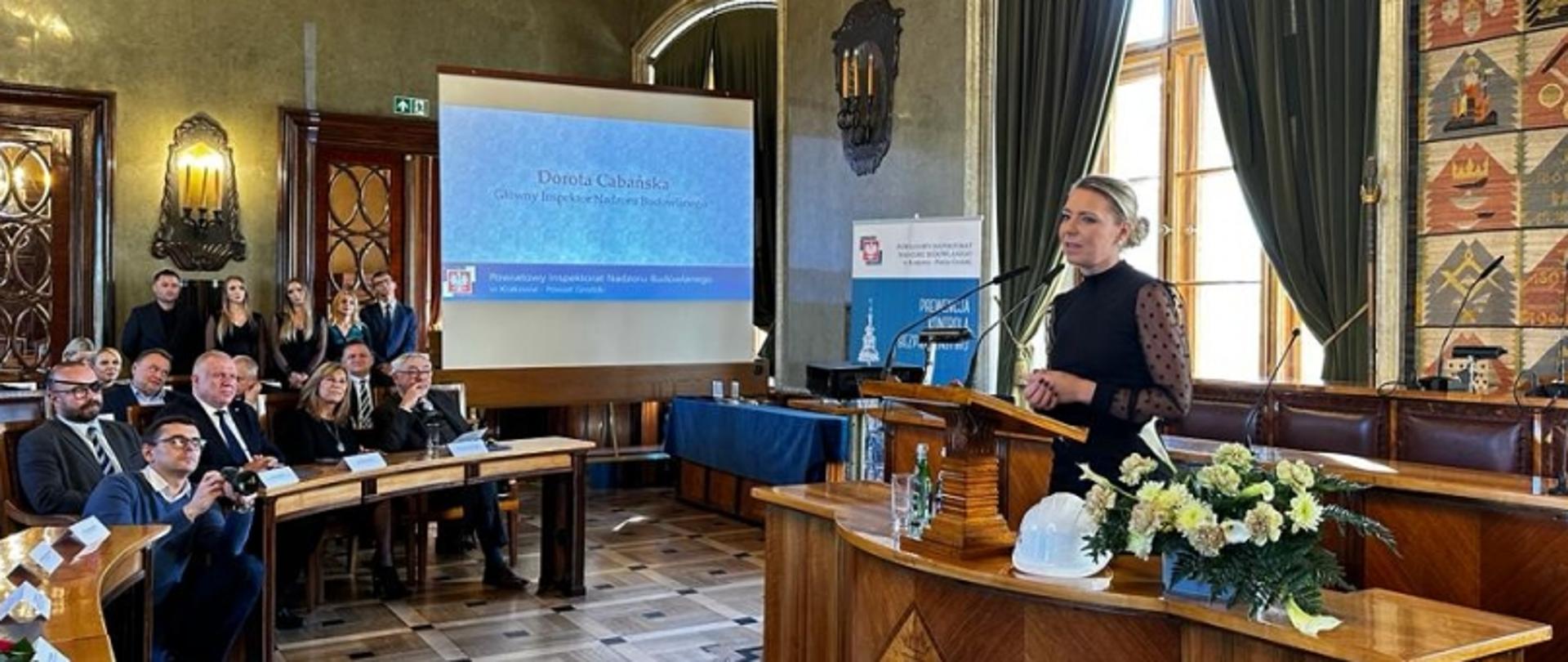 Osoba przemawiająca przy mównicy w sali konferencyjnej; na ekranie widoczna prezentacja Doroty Cabańskiej, Głównego Inspektora Nadzoru Budowlanego.