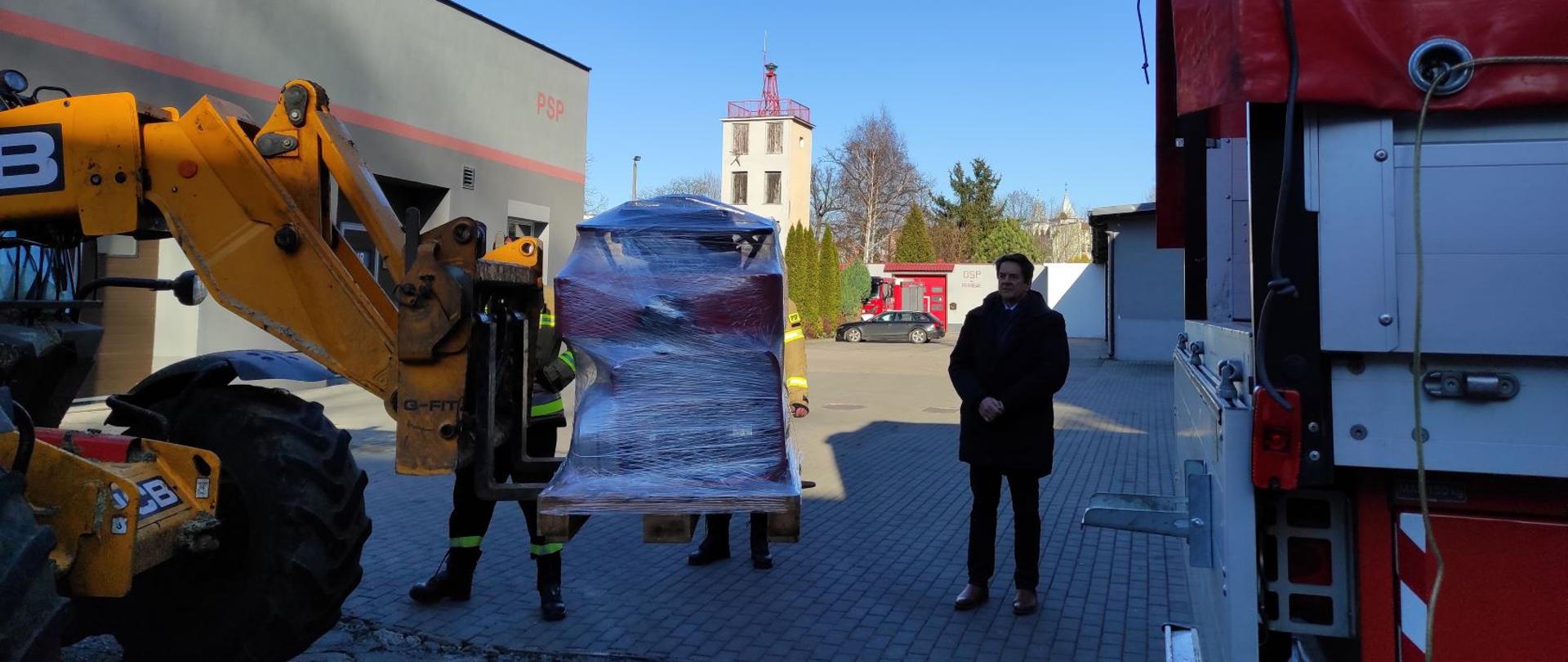 Żółta ładowarka z lewej strony wiezie na ramieniu załadowaną paletę ze sprzętem. Z prawej samochód ciężarowy