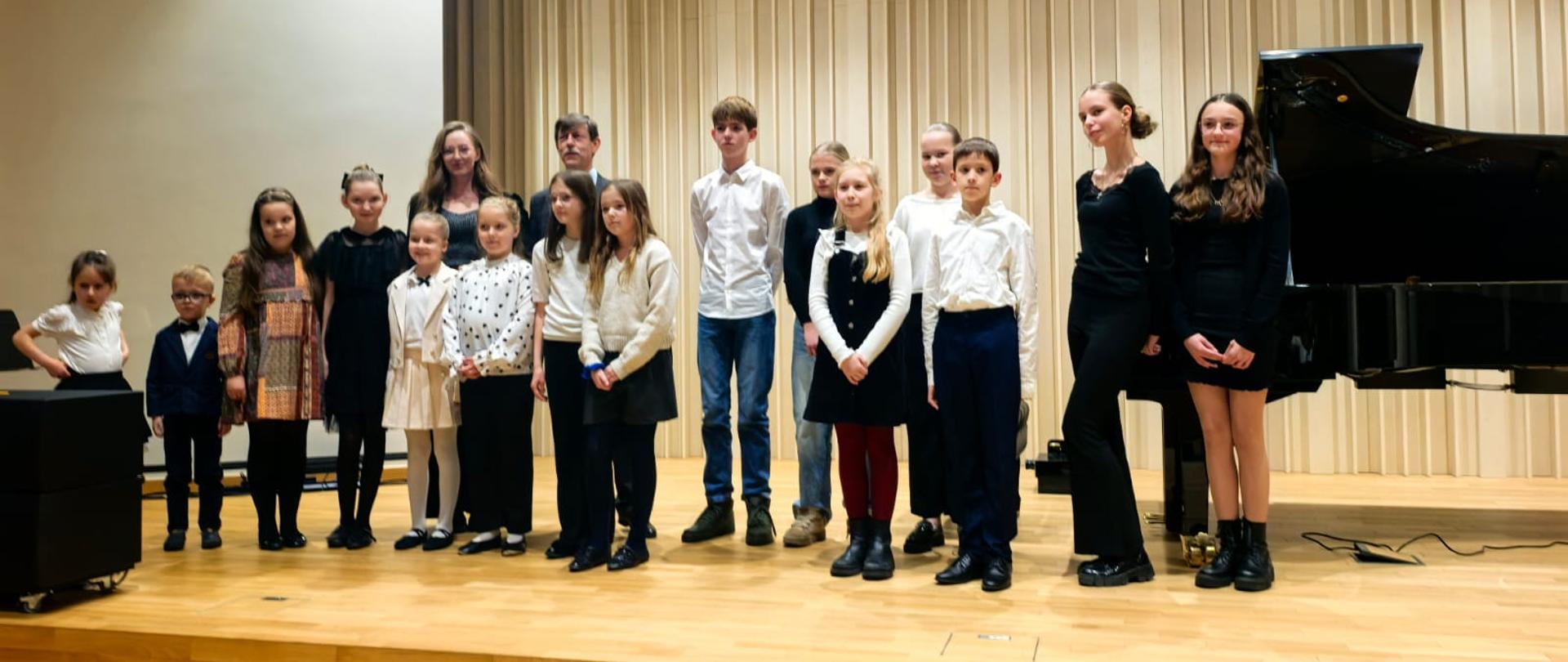 Na zdjęciu stoi grupa dzieci i nastolatków wraz z dwiema dorosłymi osobami, ustawiona w kilku rzędach na scenie sali koncertowej. Wszyscy są odświętnie ubrani i patrzą w stronę aparatu. W tle widać jasne pionowe panele akustyczne oraz fragment czarnego fortepianu po prawej stronie.