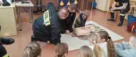 Zdjęcie przedstawia dwóch strażaków udzielających instruktarzu pierwszej pomocy dla dzieci ze szkoły w klasie.