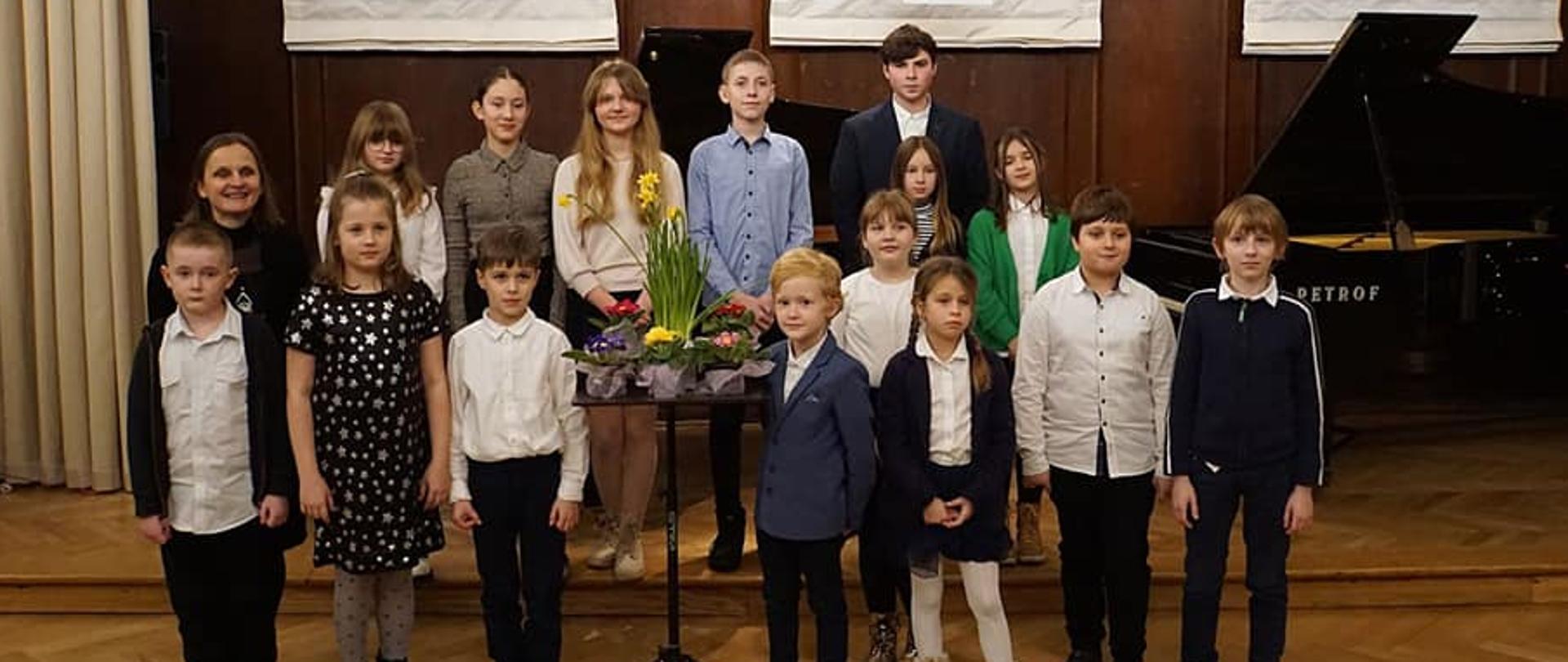 Zdjęcie przedstawia wykonawców koncertu i nauczyciela . Wszyscy stoją na środku sceny. Przed nimi wyeksponowany bukiet z kwiatów. Z tyłu widać dwa fortepiany.